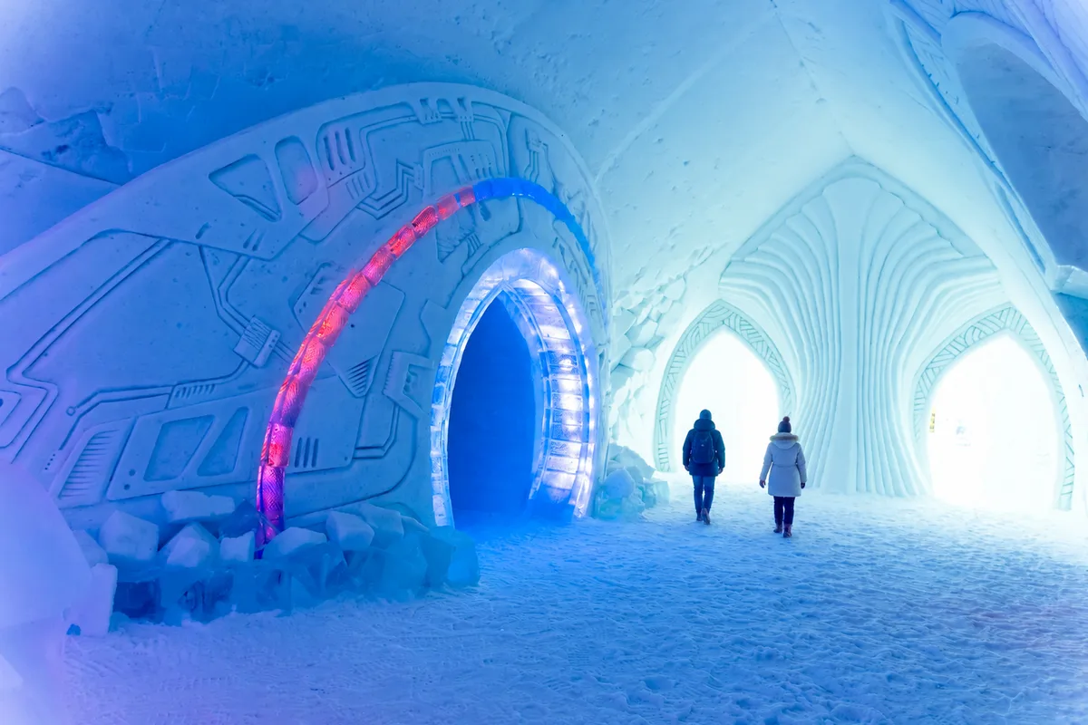 Corridoio interno dell’ICEHOTEL in Svezia con pareti e archi di ghiaccio illuminati di blu e due visitatori che camminano verso l’uscita