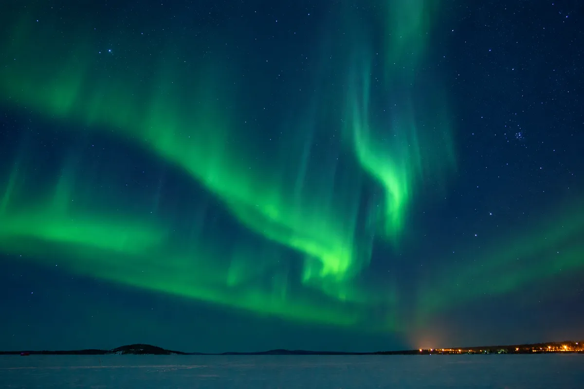 Aurora boreale verde sopra il lago ghiacciato di Inari, con le luci del villaggio lappone all’orizzonte in una notte stellata