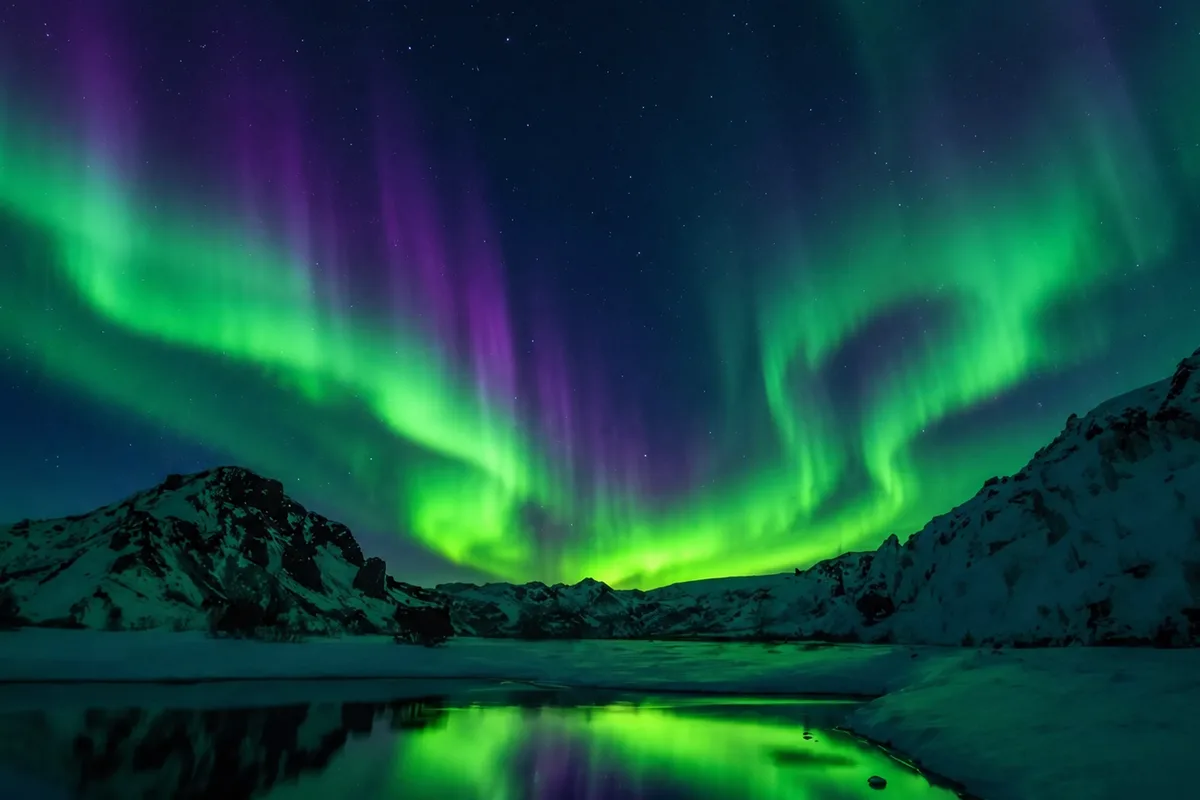 Aurora boreale verde sopra montagne innevate, riflessa in un lago in parte ghiacciato in una notte artica stellata