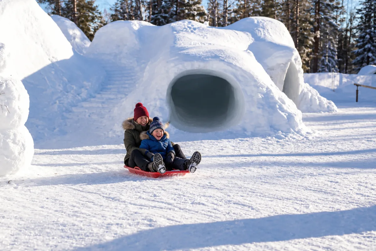 Due persone in slitta accanto a un pupazzo e un tunnel di neve allo Snowman World a Rovaniemi.